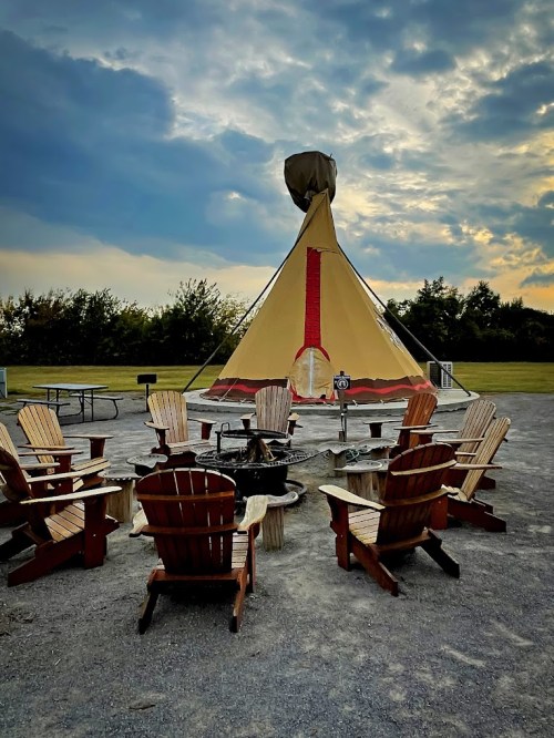 A teepee surrounded by wooden chairs and a fire pit, set against a cloudy sky.