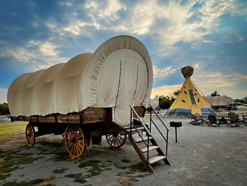 A covered wagon and a teepee are set against a cloudy sky, with outdoor seating nearby.