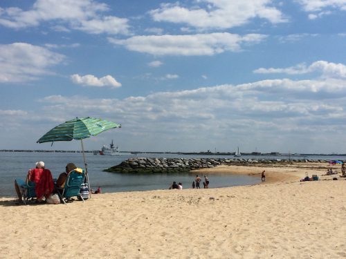 Two people relax under a striped umbrella on a sandy beach, with a calm sea and distant boats in the background.