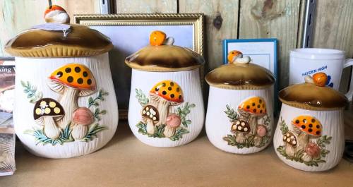 Four decorative ceramic canisters with mushroom designs, featuring brown tops and colorful polka dots, displayed on a shelf.