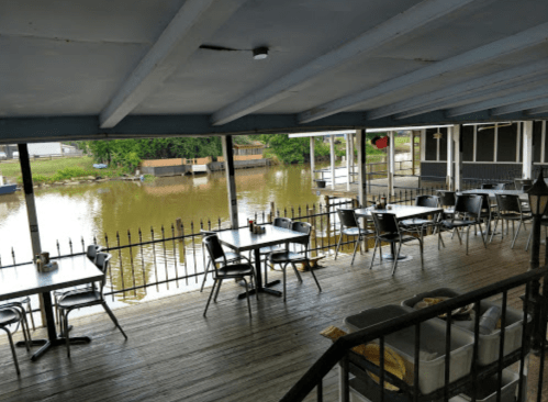 Outdoor dining area with empty tables overlooking a calm river, surrounded by greenery and wooden structures.