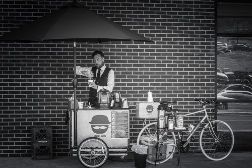 A man in a suit serves coffee from a bicycle cart under an umbrella, set against a brick wall.