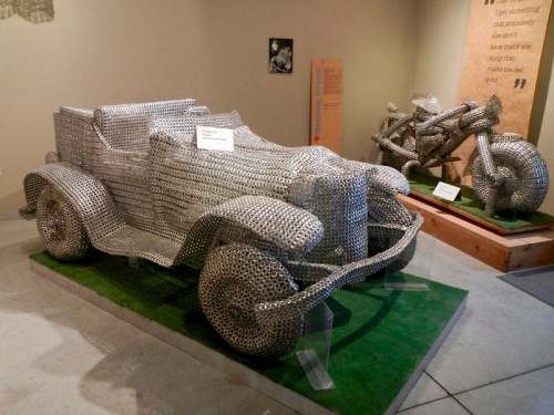 Sculptures of a vintage car and motorcycle made from metal chains, displayed in a museum setting.