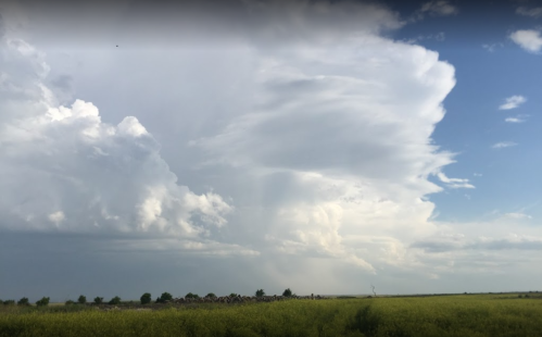 A vast sky filled with dramatic clouds, transitioning from bright blue to gray, over a grassy landscape.