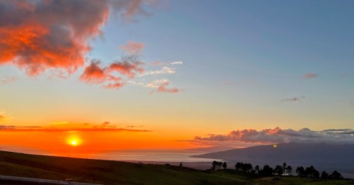 A vibrant sunset over the ocean, with colorful clouds and silhouettes of distant mountains and trees.