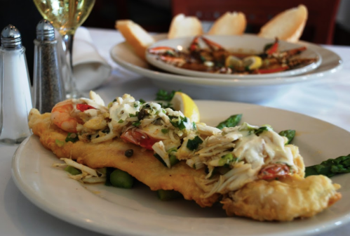 A plate of fried fish topped with crab and shrimp, garnished with lemon and asparagus, with another dish in the background.