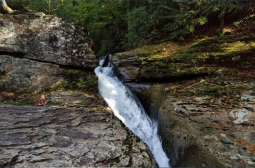 A narrow stream of water flows through a rocky landscape, surrounded by lush greenery and moss-covered stones.