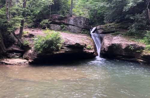 A serene waterfall cascading into a calm pool, surrounded by lush greenery and rocky terrain.