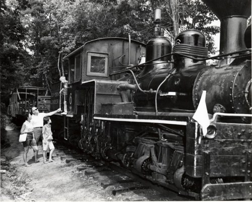 A vintage steam locomotive with passengers nearby, surrounded by trees in a black and white photograph.