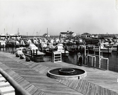 Black and white photo of a marina with boats docked, featuring a wooden boardwalk and buildings in the background.