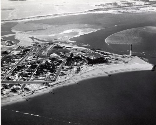Aerial view of a coastal area with a lighthouse, sandy beach, and surrounding land, captured in black and white.