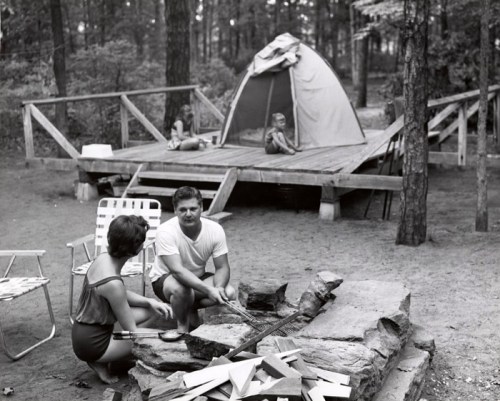 A couple sits by a campfire, while a tent and a child are visible in the background among trees.