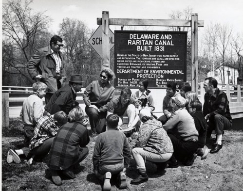 A group of children and adults gather around a historical sign about the Delaware and Raritan Canal, discussing its significance.