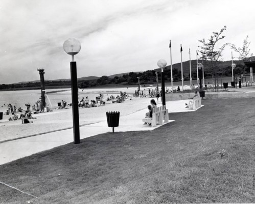 Black and white photo of a beach scene with people sunbathing, benches, and lampposts along the shore.
