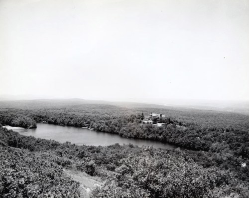 A black and white landscape showing a river surrounded by dense trees and a distant building on a hillside.