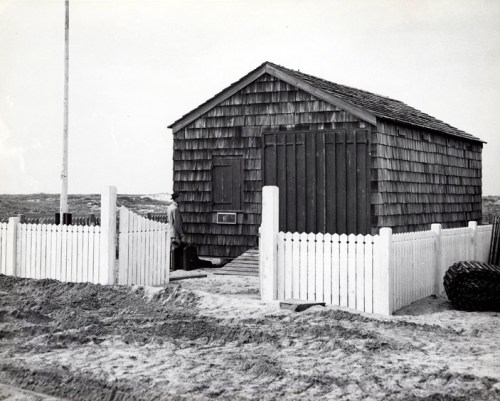 A small wooden shack with a shingled roof, surrounded by a white picket fence on a sandy beach.