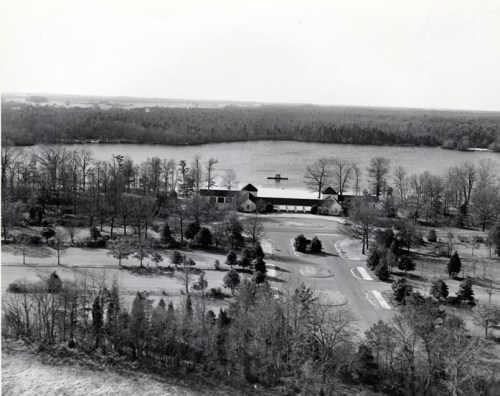 Aerial view of a lakeside property surrounded by trees and a winding road, with a calm lake in the background.