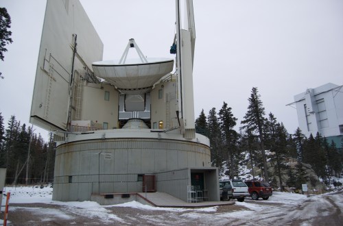 A large satellite dish on a snowy landscape, with a building structure and trees in the background.