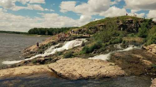 A scenic view of a rocky shoreline with small waterfalls, people exploring the area under a partly cloudy sky.