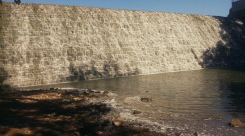 A stone dam with water cascading down into a calm pond, surrounded by trees and a clear blue sky.