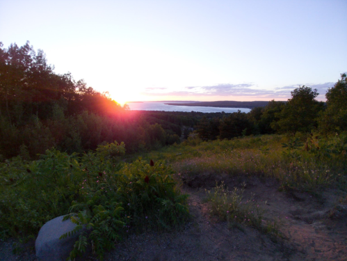 Sunset over a lush landscape, with trees and a distant body of water, casting warm colors across the sky.