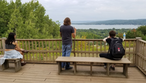 Three people on a wooden deck overlooking a scenic lake and green landscape, with cloudy skies above.