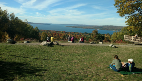 A scenic view of a lake surrounded by colorful autumn trees, with people sitting on a grassy hill.