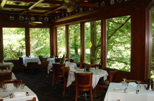 A cozy restaurant with wooden decor and large windows overlooking a green landscape, featuring neatly set tables.