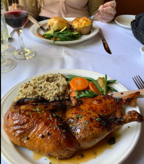 A plate with grilled chicken, rice, and vegetables, with another plate of food in the background and drinks on the table.