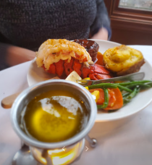 A plate featuring lobster tail, steak, baked potato, green beans, and a small dish of olive oil.