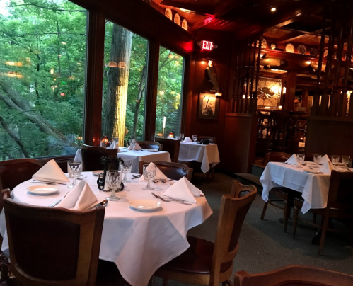 Cozy restaurant interior with wooden decor, white tablecloths, and large windows overlooking a green forest.