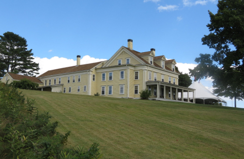 A large yellow historic house on a grassy hill, with a tent set up nearby under a blue sky.