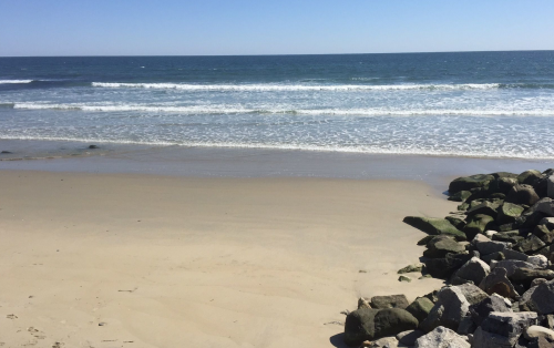 A serene beach scene with gentle waves lapping at the shore and rocky formations along the sand. Clear blue sky above.