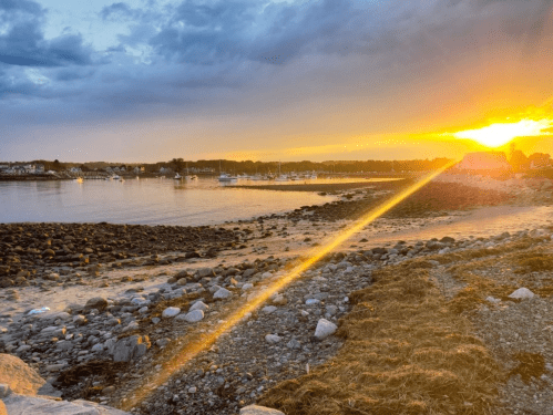Sunset over a rocky shoreline with boats in the water, casting a golden light across the scene.