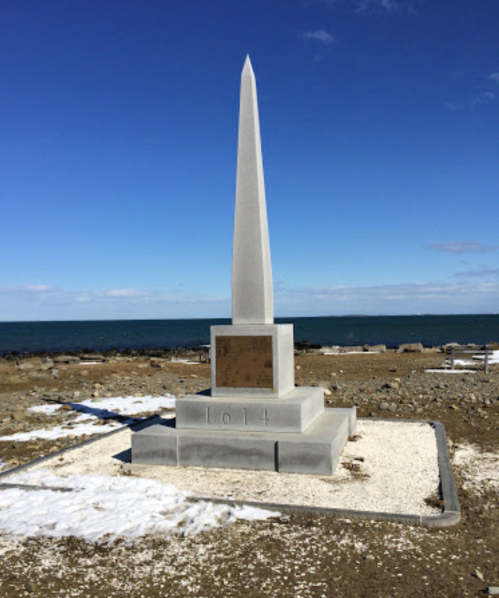 A tall stone obelisk monument on a rocky shore, with a clear blue sky and ocean in the background. Snow is visible nearby.