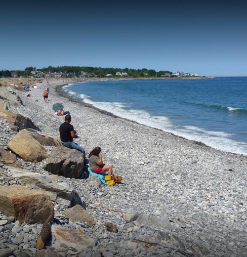 A rocky beach scene with people sitting and relaxing by the water on a sunny day.