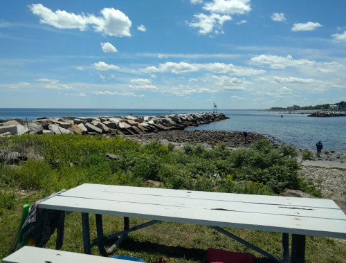 A scenic view of a rocky shoreline with a lighthouse in the distance, under a bright blue sky with fluffy clouds.