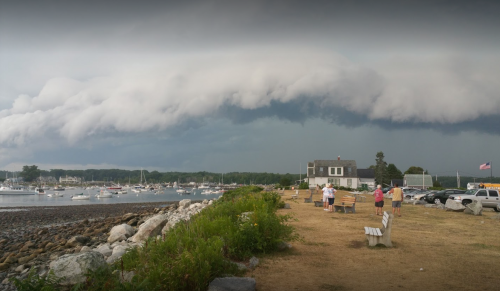 A stormy sky looms over a harbor with boats, while people gather on the shore near a building and parked cars.