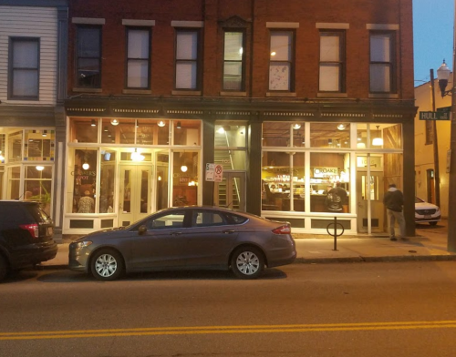 A street view of a brick building with large windows, featuring a restaurant and parked cars in front.