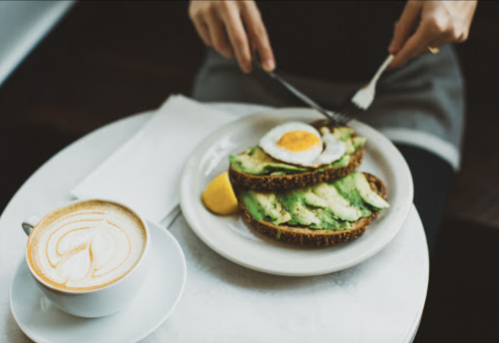 A person slices an avocado toast topped with a poached egg, alongside a cup of latte on a table.