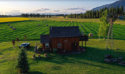 A rustic wooden house surrounded by green fields and hay bales, with a windmill in the background and mountains nearby.