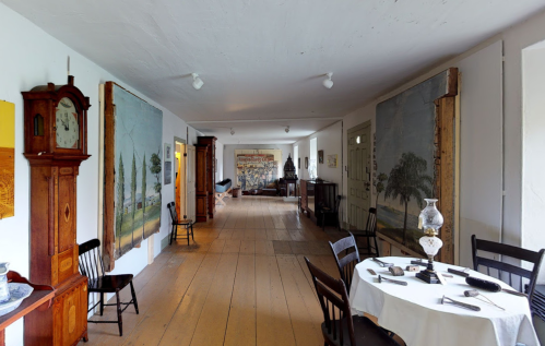 A spacious room with wooden floors, vintage furniture, and large murals on the walls, featuring a dining table and chairs.