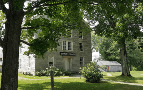 A historic stone building with large windows, surrounded by trees and grass, featuring a sign for the Historical Society.