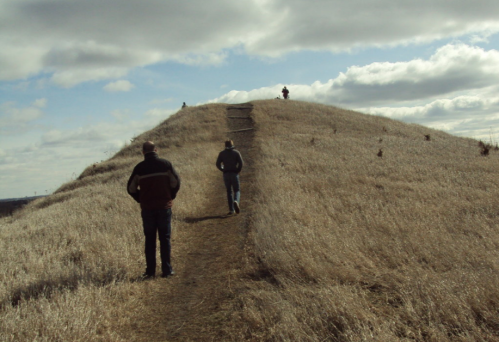 Two people walking on a grassy hill under a cloudy sky, with a third person visible at the top of the hill.