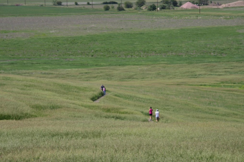 Two people walk along a path through a lush green field, with rolling hills and distant structures in the background.