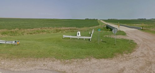 A rural intersection with a gravel road and a grassy area, surrounded by fields under a clear sky.