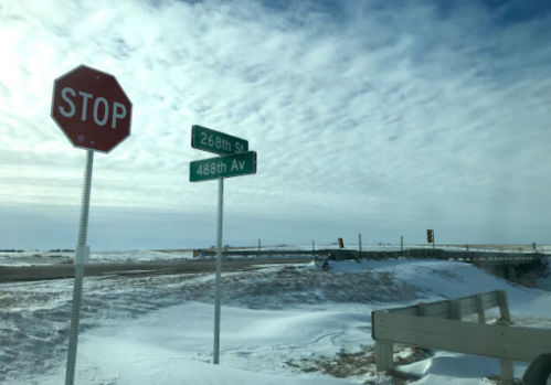 A stop sign at the intersection of 268th Street and 488th Avenue, with a snowy landscape and cloudy sky in the background.
