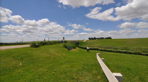 A grassy area with people working near a road under a blue sky with fluffy clouds and fields in the background.