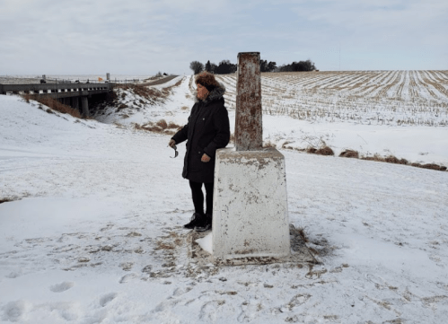 A person stands beside a weathered concrete marker in a snowy landscape, with a bridge and fields in the background.