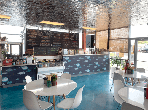 Bright, modern café interior with blue floors, white tables, and a menu board on the wall. Plants add a touch of greenery.